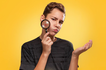 Wall Mural - Cosmetology and acne. A disgruntled teenage boy in a black t-shirt with pimples on his face, holds a magnifying glass to his cheek, and spreads his hand to the side. Yellow background. Copy space