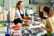 © JackF - Portrait of friendly female fishmonger showing raw European bass to woman behind counter of seafood store