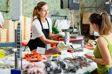 Portrait Of Friendly Female Fishmonger Showing Raw European Bass To Woman Behind Counter Of Seafood Store