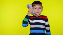 Rich Little Child Boy In A Colorful Sweater Puts A Lot Of Money To His Ear Imitating A Mobile Phone Then The Dollars Fall Down On A Yellow Background With Copy Space. Place For Text Or Product