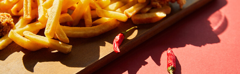 Wall Mural - spicy french fries served on wooden cutting board with chili peppers in sunlight, panoramic shot