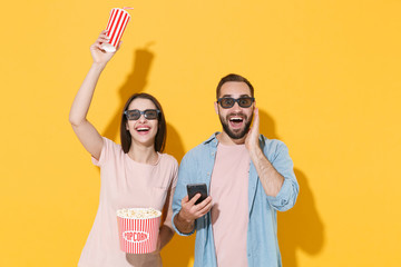 Wall Mural - Excited young couple friends guy girl in 3d glasses isolated on yellow background. People in cinema concept. Watching movie film, hold bucket of popcorn, cup of soda mobile phone put hand on cheek.