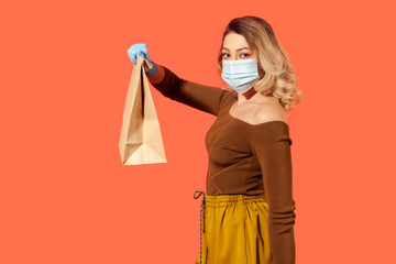 Side view woman in protective mask giving paper bag and looking at camera, advertise of food purchase delivery in coronavirus self-isolation, online order. studio shot isolated on red background