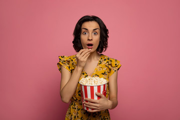 Watching movies. Close-up photo of a young surprised lady, who is eating popcorn and looking in the camera.