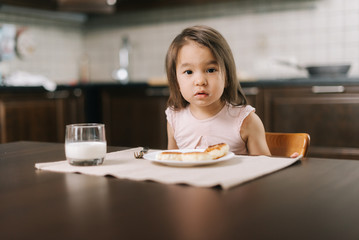 Beautiful little two-year-old girl is waiting for the meal to begin, looking at the camera.. Cheesecakes on the table in front of the child and a glass of milk for breakfast.