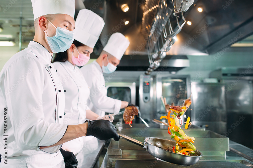 Chefs in protective masks and gloves prepare food in the kitchen of a restaurant or hotel. - obrazy, fototapety, plakaty 