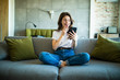 Portrait of a surprised woman holding a phone looking at you sitting on a sofa in the living room