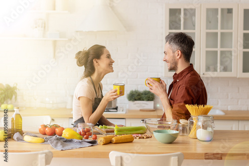 Happy loving couple chatting during coffee break standing at home kitchen. Handsome husband and beautiful wife together. Woman in apron cook fresh vegetable salad. Man entertain beloved soulmate