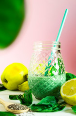 On a light wooden table there is a glass jar with a green vitamin smoothie of fruits and vegetables. Next to the smoothie bowl are apples, bananas, a lemon and spinach leaves.