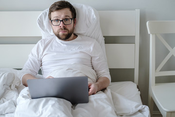 Young, bewildered man in white pajamas remotely works with a laptop while sitting in bed. Modern lifestyle.