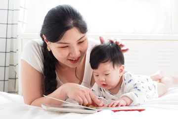 Warm moment of Asian mother playing and teaching her cute newborn baby with cartoon picture book in the bedroom at home. Storytelling, happy family, mother and baby lifestyle and pure love concept