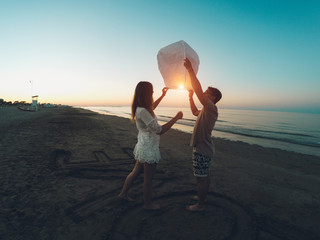 Couple of lovers lighting sky lantern on the beach at sunset - Young people celebrating their relation anniversary in summer vacation - Love and travel concept - Main focus on woman's head