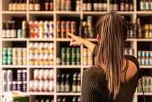 A Young Woman Pointing At Some Canned Beers In A Liquor Shop And Choosing. Beers From Trendy Small Breweries Displayed On Shelfs. Bokeh Background