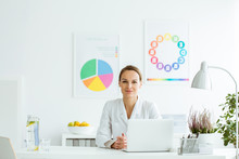 Beautiful Doctor Sitting At The Desk In Her White Office With Colorful Boards In The Background