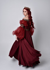 Wall Mural - Portrait of a beautiful woman with red hair wearing  a  flowing Burgundy fantasy gown and golden crown.  full length standing pose, isolated against a studio background
