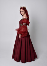 Portrait Of A Beautiful Woman With Red Hair Wearing  A  Flowing Burgundy Fantasy Gown And Golden Crown.  Full Length Standing Pose, Isolated Against A Studio Background
