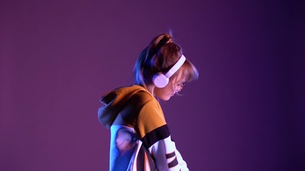 Wall Mural - Stylish gen z teen girl wear headphones listening dj pop music playlist dancing on violet neon light color studio background. 20s hipster fashion woman in hoodie glasses at cool party, slow motion.