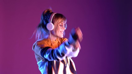 Wall Mural - Gen z teen girl model wear headphones listening dj pop music playlist dancing on purple violet neon light color studio background. Stylish 20s hipster fashion woman in hoodie sunglasses at cool party.