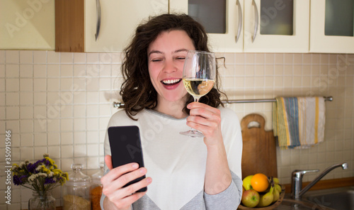 Happy young smiling caucasian woman making video call with smartphone at kitchen. Best friends drinking white wine and toasting. Video conference party online meeting with family. Stay home.