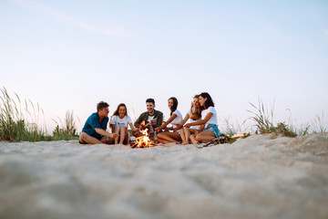 group of young friends sitting on beach and fry sausages. one man is playing guitar. summer holidays