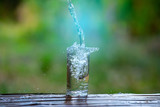 Fototapeta Łazienka - Drink water pouring in to glass over sunlight and natural green background.Water splash  in glass Select focus blurred background.