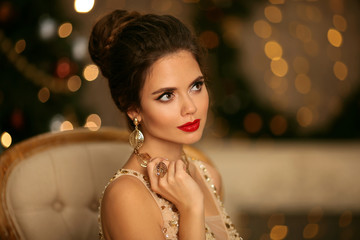 Wall Mural - Beauty portrait of elegant woman with wedding hairstyle and makeup. Beautiful brunette girl with golden jewelry in prom dress sitting on modern chair over bokeh lights xmas decorations.