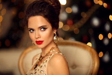 Wall Mural - Luxurious portrait of elegant woman with wedding hairstyle and makeup. Beautiful brunette girl with golden jewelry in prom dress sitting on modern chair over bokeh lights xmas decorations.