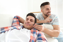 Young Man Drawing Mustache On Face Of Sleeping Friend Indoors. April Fool's Day