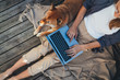 © iana_kolesnikova - Top-down image of young woman working remotely on laptop computer on home wooden terrace typing text message on keyboard her best friend dog lies next to her sleeping, Working From Home Lifestyle