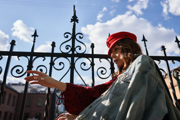 A modern young woman in red dress, denim jacket and red cap, posing on the street of the city with sky in background. Beauty, street fashion. Hand motions.