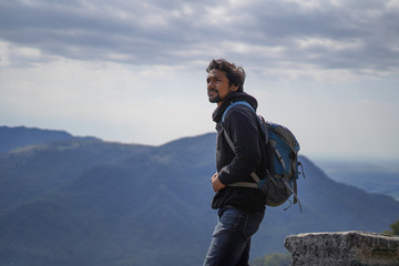 young handsome indian boy standing on the top of the mountain with a bag pack while looking upwards 