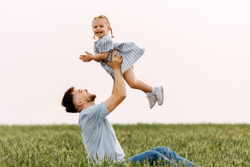 Young man and little girl playing on a summer day, in a green field, outdoors. Father holding his daughter up to the sky, sitting on the ground.