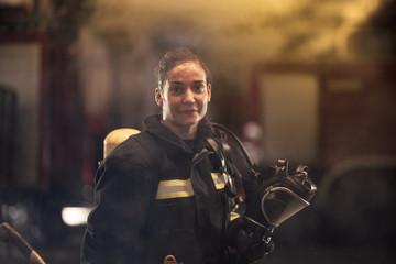 Wall Mural - female firefighter portrait wearing full equipment and emergency rescue equipment. taking off oxygen mask after successful intervention. smoke and fire trucks in the background.