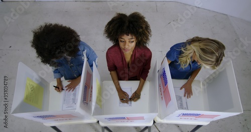 LS Wide overhead view three young women with mixed-race woman in center, looking thoughtful as she fills in ballot at voting booths in polling station.