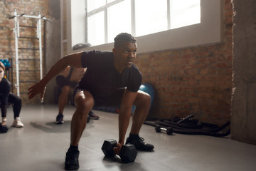 Wall Mural - Train hard, live better. Full length shot of athletic man lifting dumbbell while having workout at industrial gym. Group training, teamwork concept