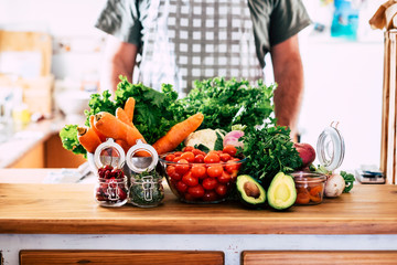 Home kitchen wooden table full of fresh and coloured vegetables ready to be cooked and prepared for healthy nutrition lunch or dinner - unrecognizable chef man people in background