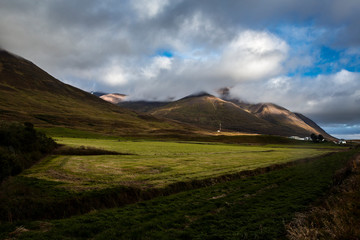 landscape in iceland with is a green meadow. the sky is full of clouds and we can see torrents of water falling in the middle of the sun's rays