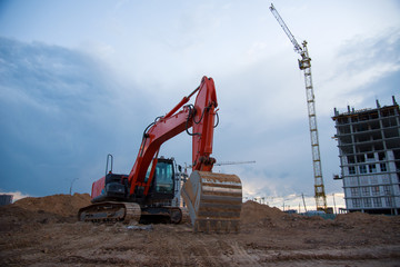 Excavator working at construction site on earthworks on evening sunset. Backhoe digs ground for roadwork. Tower cranes in action construction of a new multi-storey buildings in night