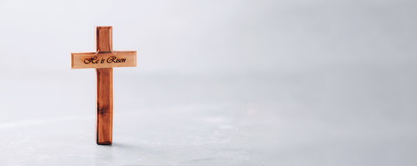 Wooden cross with text He is risen on grey background. Reminder of Jesus sacrifice and Christ resurrection. Easter passover. Eucharist concept. Christianity symbol and faith