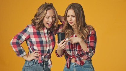 Wall Mural - Young optimistic pretty girls isolated over yellow wall background using mobile phone