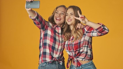 Sticker - Young positive happy girls isolated over yellow wall background take a selfie by mobile phone