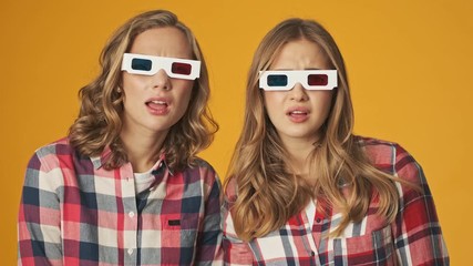 Poster - Young shocked scared girls friends sisters isolated over yellow wall background watch film in 3d glasses