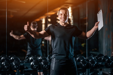 young cheerful personal trainer in gym and with clipboard and stopwatch