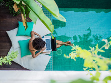 Bird View Of A Remote Online Working Digital Nomad Women With Curly Hair And Laptop Sitting At A Sunny Turquoise Water Pool Having Feet In The Water Surrounded By Cushions And Plants In The Foreground