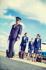 Wall Mural - Portrait of mature pilot walking with three young beautiful flight attendants against airplane in airport