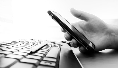 Man's hand holds a smartphone over a black computer keyboard. Heavily blurred background. Black and white image. Copy space.