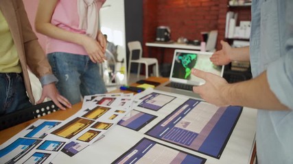Wall Mural - Hands of unrecognizable business team of three discussing printouts with website interface templates for different devices standing around desk in office, young man gesturing while explaining ideas