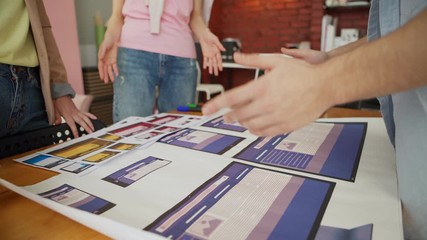 Wall Mural - Closeup panning shot of startup business team of three people discussing printouts with app interface templates standing around desk in office, male designer gesturing while explaining ideas