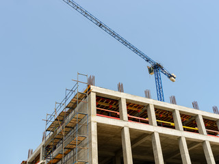 Concrete building under construction and blue tower crane with cargo on a sunny day. Modern construction machinery against blue sky. Scaffolds on the wall.