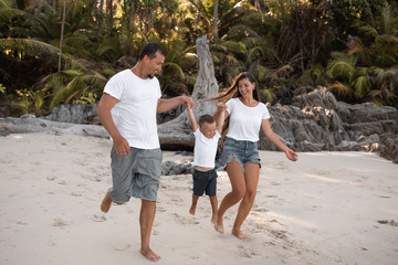 Wall Mural - Happy smile european family have rest and run on white sand beach. Father, long black chestnut hair mother, blond baby boy. white cotton clothes. boho dress. t-shorts.Thailand. Aquamarine crystal sea